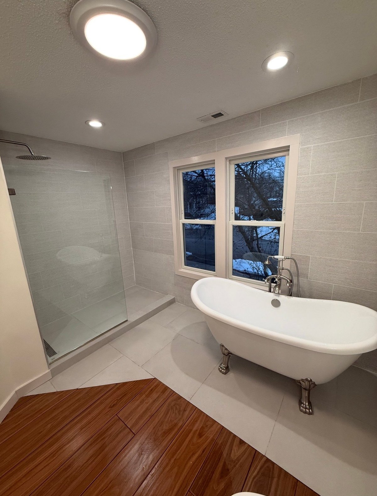 Modern bathroom with white subway tile walls, freestanding white tub, glass shower enclosure, wood-look floor, and large windows