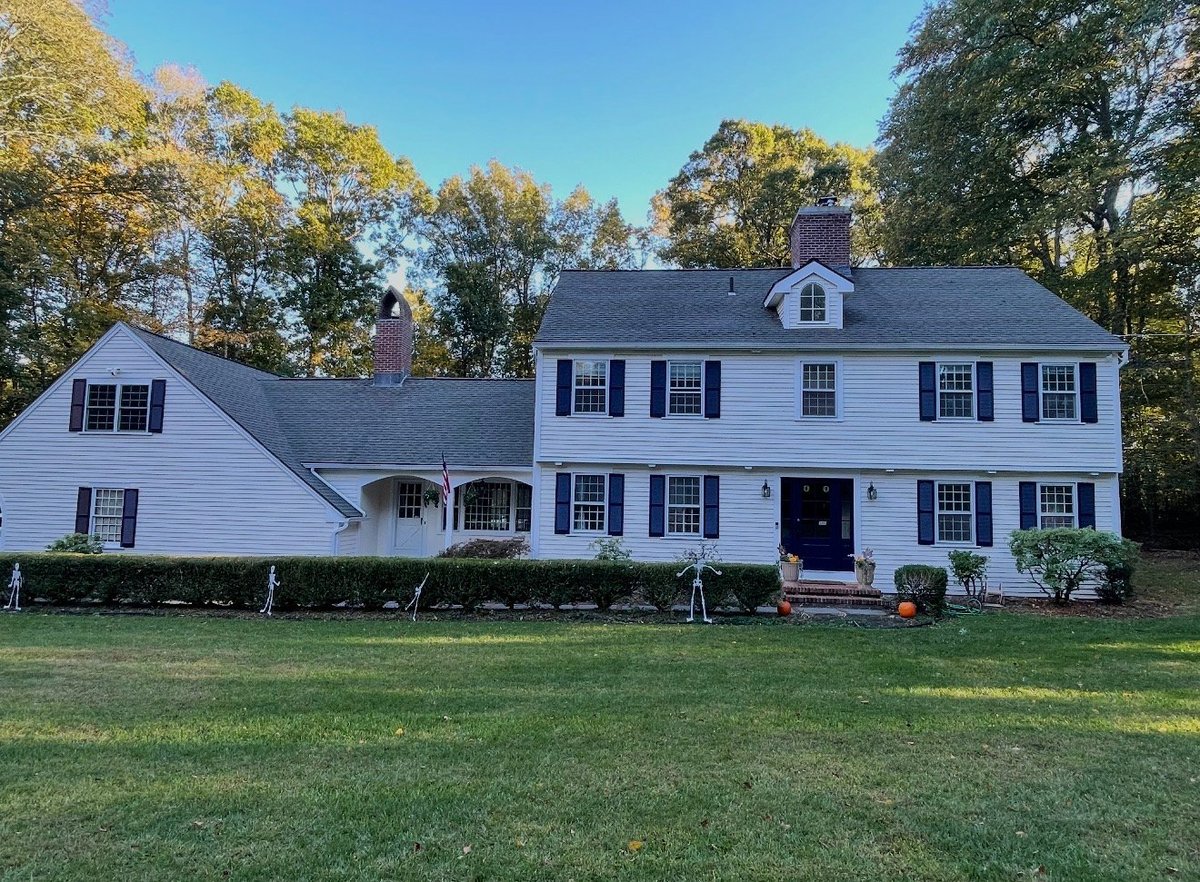 White two-story colonial home with dark shutters, front porch, and attached garage on manicured lawn with autumn trees